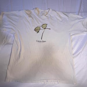 May include: White t-shirt featuring a graphic of sunglasses with a brown frame and lenses. The shirt has the Calvin Klein logo printed in black. The shirt is slightly wrinkled and appears to be vintage.