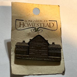 May include: A bronze-toned metal pin shaped like a barn, mounted on a rectangular beige card. The pin features the text CRAWFORD BARN at the base. The card displays the text LONGABERGER HOMESTEAD at the top with a small leaf graphic below it.