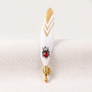May include: A white feather brooch with gold chevron accents and a black spider with a red gem. The brooch has a gold-colored pin backing.