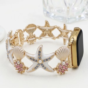 May include: Gold-toned Apple Watch band featuring white starfish and seashell charms. The band has blue and pink crystal accents and a gold clasp. The watch face is black.