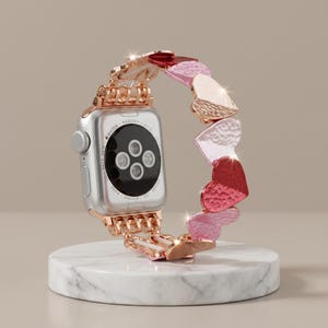 May include: A silver smartwatch with a black circular sensor array on the back, attached to a decorative metal watch band composed of interconnected heart-shaped links in alternating shades of rose gold, light pink, and deep red with a hammered texture.