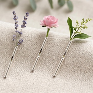 Könnte beinhalten: Drei silberne Einzelvasen, jede mit einer anderen Blume oder einem Zweig. Eine Vase enthält Lavendel, eine andere eine rosa Rose und die dritte grüne Blätter und winzige weiße Blüten. Die Vasen stehen auf einem neutralfarbenen Stoff.