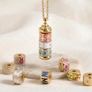 Puede incluir: Collar dorado con un colgante cilíndrico con bandas de esmalte apiladas y coloridas con números y símbolos. Varias cuentas doradas con letras y piedras preciosas están dispersas.