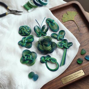 May include: An assortment of handcrafted, decorative knots in shades of green and blue, arranged on a white cloth. The knots vary in shape, including floral and butterfly designs. A pair of pliers and spools of thread are visible in the background.