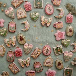 Puede incluir: Una colección de colgantes de cerámica pintados a mano con formas caprichosas, incluyendo corazones, lunas, mariposas y sellos. Los colgantes presentan diseños florales y de inspiración natural en colores pastel, con el texto "COZY VIBES ONLY" en algunos.