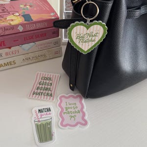 May include: A black handbag with a heart-shaped keychain that reads "But First, Matcha." Several stickers with matcha-themed phrases and a stack of books are also visible. The stickers say "Cool Girls Drink Matcha," "Matcha Girlie," and "I love you so matcha."