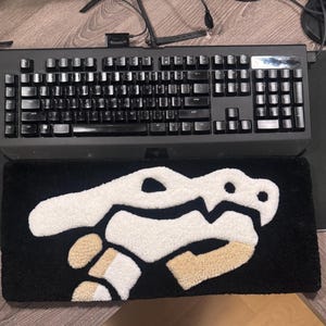 Op de afbeelding: Zwart toetsenbord bovenop een zwarte polssteun met een Bowser-ontwerp. De polssteun heeft een zwart oppervlak met witte en beige details. Het toetsenbord en de polssteun liggen op een houten oppervlak.