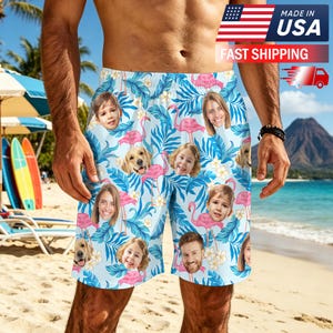 May include: Light blue swim trunks featuring a repeating pattern of pink flamingos, white tropical flowers, blue palm leaves, and various photographic portraits of children and a golden retriever dog printed across the fabric.