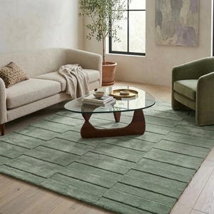 May include: A living room scene featuring a light green rug with a geometric pattern. A cream-colored sofa and a green armchair are arranged around a glass-topped coffee table with a wooden base. An olive tree in a terracotta pot adds a natural element.