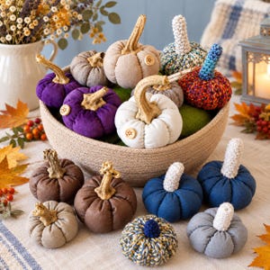 Peut inclure: Une collection de citrouilles décoratives en tissu de différentes couleurs, notamment violet, blanc, marron, bleu et gris, avec des tiges texturées et de petits boutons en bois. Certaines sont en tissu uni, d'autres sont crochetées.