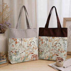 Op de afbeelding: Twee tote bags met een botanische print in gedempte oranje, groene en bruine bloemmotieven op een crèmekleurige achtergrond. De linker tas heeft lichtgrijze hengsels en bovenrand, de rechter tas heeft donkerbruine hengsels en bovenrand.