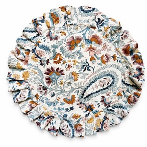 May include: A round, floral-patterned placemat with a ruffled edge. The design features a paisley center and a repeating pattern of flowers and leaves in shades of blue, brown, and pink on a white background.