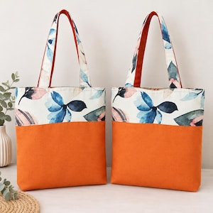 May include: Two identical tote bags featuring a solid orange bottom section and a white upper section decorated with a watercolor-style floral pattern in shades of blue, pink, and dark green. Each bag has two matching fabric handles.