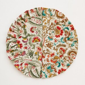 May include: A circular fabric placemat featuring a ruffled edge and a cream-colored base with a dense floral and paisley pattern in shades of red, green, yellow, and teal. The design is split into two distinct halves with different botanical prints.
