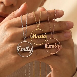 Peut inclure: Trois colliers personnalisés avec des noms en argent, or et or rose. Chaque collier présente un pendentif circulaire avec un nom en écriture cursive. Les noms sont "Emily" et "Maria". Les colliers sont présentés sur un fond neutre.