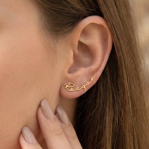 Puede incluir: Pendientes trepadores dorados que deletrean el nombre "Raegan" en escritura cursiva. Los pendientes se llevan en una oreja, mostrando el diseño delicado y el toque personalizado. Joyas de estilo minimalista.