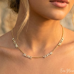 Op de afbeelding: Een goudkleurige ketting met de namen "Evelynn", "Blake", "Sophie" en "Nicole" in afzonderlijke letters. De ketting wordt om de nek gedragen. De achtergrond is wazig, met een warm, neutraal kleurenpalet.