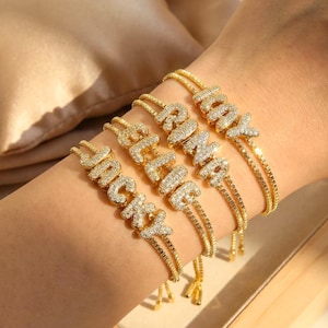 Peut inclure: Bracelets dorés épellent les noms "JACKY", "ALEX" et "RAMAY". Chaque bracelet est orné de pierres précieuses claires et scintillantes incrustées dans les lettres. Les bracelets sont portés sur un poignet, avec un fond doux et neutre.