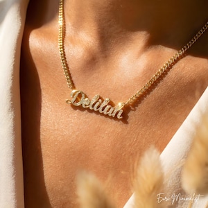 Könnte beinhalten: Eine goldfarbene Halskette mit dem Namen "Delilah" in Schreibschrift, verziert mit kleinen, glitzernden Steinen. Die Halskette, mit einer Panzerkette, wird am Hals einer Person gezeigt. Das Design ist elegant und minimalistisch.