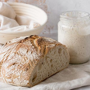 Può includere: Una pagnotta di pane a lievitazione naturale dalla crosta dorata con la parte superiore incisa, accanto a un barattolo di vetro trasparente pieno di lievito madre attivo e un cestino rotondo color crema foderato con un panno chiaro.