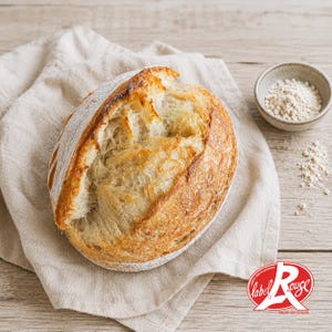 Könnte beinhalten: Ein runder Sauerteiglaib mit einer goldbraunen, knusprigen Kruste und einem tiefen Schnitt auf der Oberseite. Das Brot ist mit weißem Mehl bestäubt und liegt auf einem hellbeigen Leinentuch, begleitet von einer kleinen Schale mit Mehl.