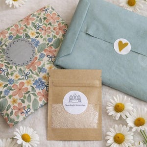 Peut inclure: Vue aérienne d'un paquet de levain, d'un carnet à motifs floraux et d'un colis emballé avec un autocollant en forme de cœur. Des marguerites blanches à cœur jaune sont dispersées autour des objets, créant une esthétique naturelle et accueillante.
