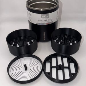 May include: Black and silver insulated food jar with the text "FOOD JAR 32oz" and several black and white components. The components include two black containers with a grid design, and two white and black lids.