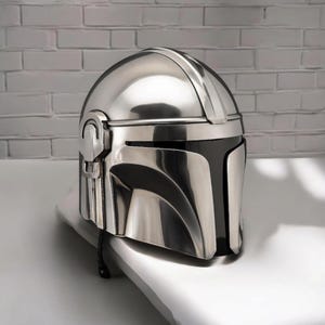 Puede incluir: Un casco plateado con una visera negra, que recuerda a un casco mandaloriano. El casco tiene una superficie lisa y reflectante, colocado sobre una superficie blanca y una pared de ladrillos grises. El dise&ntilde;o presenta l&iacute;neas y curvas distintivas.