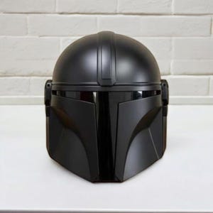 May include: A black Mandalorian helmet with a rounded crown and a distinctive angular visor. The helmet's design is sleek and modern, with a vertical ridge and side attachments. This collectible item is a replica of the iconic helmet.