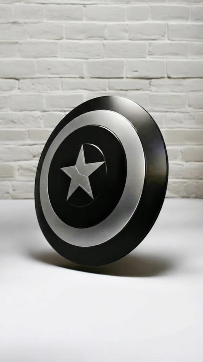 May include: A black and silver shield with a white star in the center. The shield has a circular design with a silver outer ring and a black center. The shield is leaning against a white brick wall.