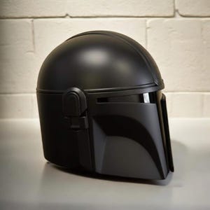 May include: A black Mandalorian helmet with a rounded crown and a visor. The helmet has a T-shaped visor and angular cheek plates. The helmet is a replica of the iconic helmet from the Star Wars series.