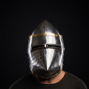 May include: A polished silver medieval helmet with a pointed top and a face guard featuring eye slits and ventilation holes. The helmet has a gold-colored band around the top and is riveted. The background is black.