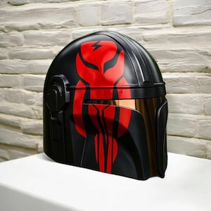 May include: A black helmet with a red graphic design. The helmet has a gold-tinted visor. The red design features a lightning bolt and vertical lines. The helmet is displayed against a white brick wall.