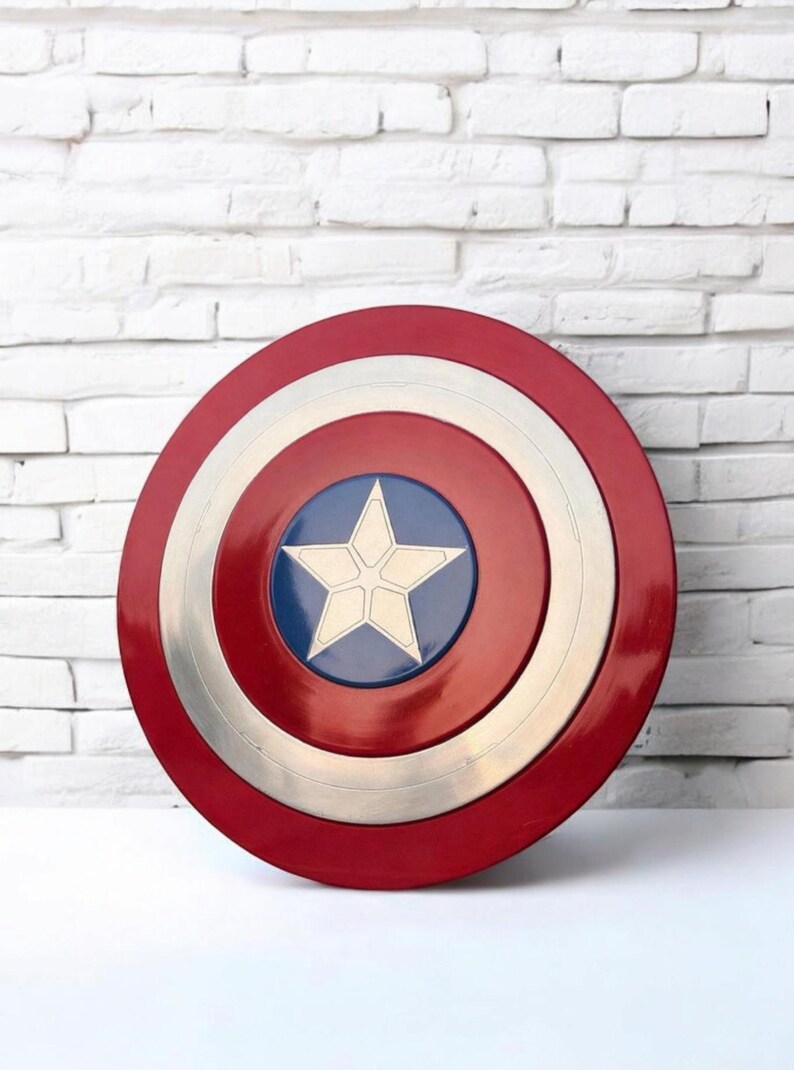 May include: A red, white, and blue Captain America shield. The shield has a red outer ring, a white inner ring, and a silver ring in the middle. The center features a blue circle with a white star. The shield is a prop or collectible.