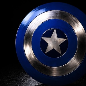 Op de afbeelding: Een blauw en zilver schild met een zilveren ster in het midden. Het schild heeft een cirkelvormig ontwerp met concentrische ringen in blauw en zilver. De ster staat in het midden van het schild. Dit is een rekwisiet of verzamelobject.