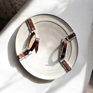 May include: A round, off-white metal plate with riveted brown leather straps. The plate has a silver rim and a central, slightly recessed area. The leather straps are secured with metal rivets and buckles, creating a decorative and functional design.