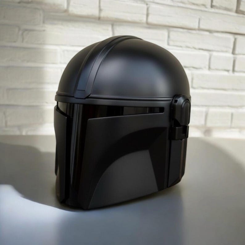 May include: A black Mandalorian helmet with a smooth, matte finish. The helmet has a rounded top and a distinctive visor, with detailed side features. The design is inspired by the Star Wars series, with a sleek and modern aesthetic. The helmet is a replica.