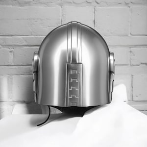 Puede incluir: Un casco plateado brillante con una parte superior redondeada y ranuras verticales. El casco presenta un acabado liso y reflectante y una zona de visera oscura. El dise&ntilde;o incorpora elementos rectangulares y curvos, d&aacute;ndole un aspecto futurista o retro.