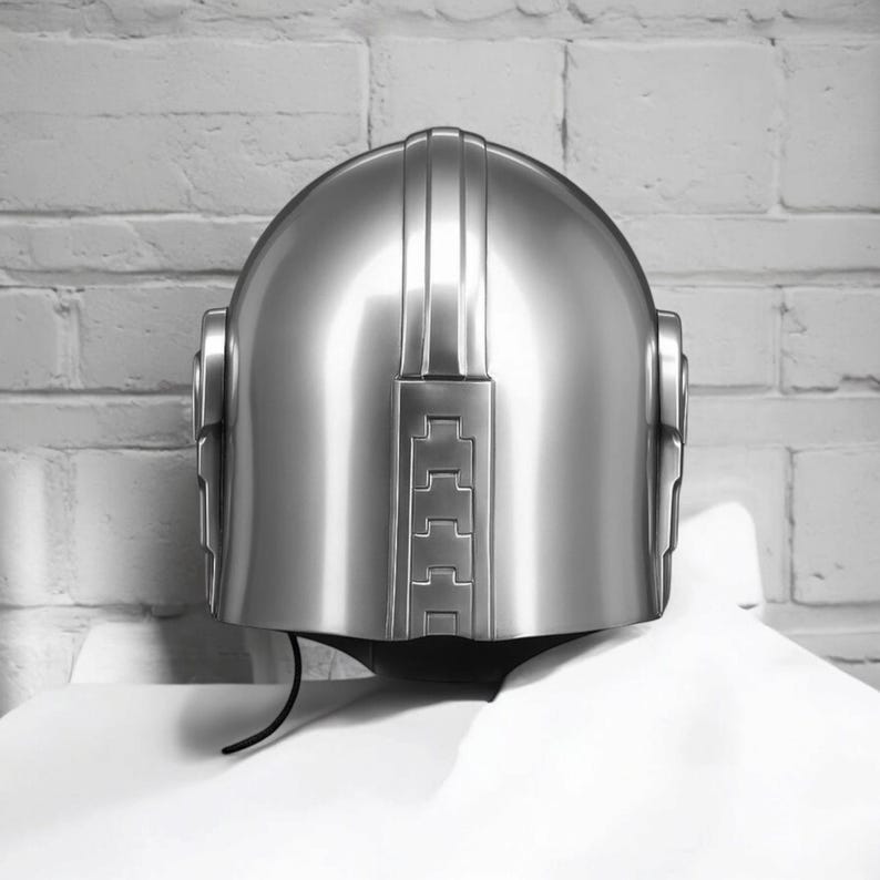 Puede incluir: Un casco plateado brillante con una parte superior redondeada y ranuras verticales. El casco presenta un acabado liso y reflectante y una zona de visera oscura. El dise&ntilde;o incorpora elementos rectangulares y curvos, d&aacute;ndole un aspecto futurista o retro.