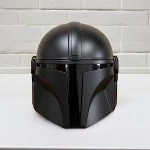 May include: A black Mandalorian helmet, inspired by the Star Wars series, is shown. The helmet has a rounded crown, a T-shaped visor, and angled cheek plates. It is set against a white brick wall. The helmet is a replica.