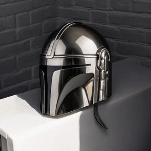 Puede incluir: Un casco plateado met&aacute;lico de Mandaloriano, con una superficie reflectante. El casco tiene una visera en forma de T y detalles laterales. Un art&iacute;culo de colecci&oacute;n.