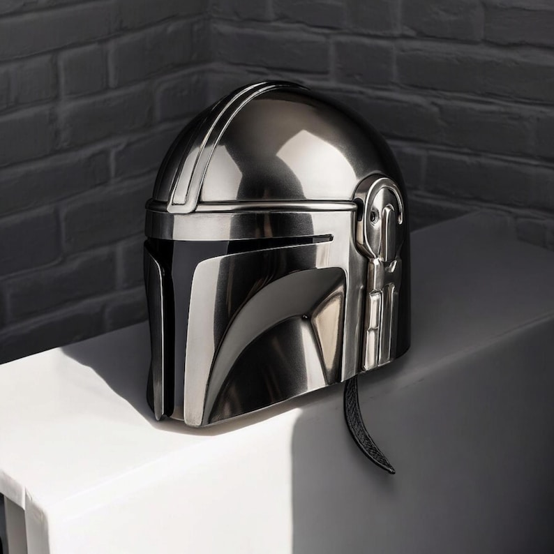 Puede incluir: Un casco plateado met&aacute;lico de Mandaloriano, con una superficie reflectante. El casco tiene una visera en forma de T y detalles laterales. Un art&iacute;culo de colecci&oacute;n.
