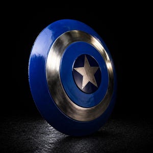 Op de afbeelding: Een blauw en zilver schild met een centrale zilveren ster. Het schild heeft een cirkelvormig ontwerp met een blauw centrum, een zilveren ring en een blauwe buitenrand. Het schild staat tegen een zwarte achtergrond.