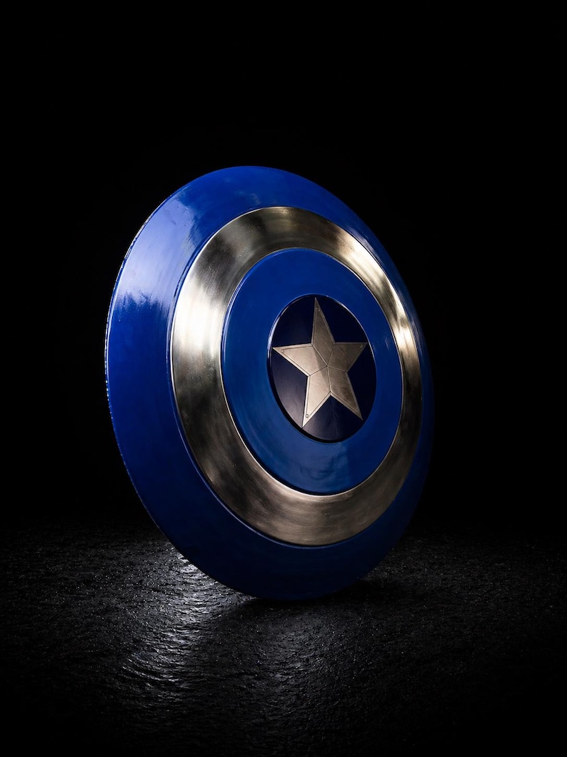 Op de afbeelding: Een blauw en zilver schild met een centrale zilveren ster. Het schild heeft een cirkelvormig ontwerp met een blauw centrum, een zilveren ring en een blauwe buitenrand. Het schild staat tegen een zwarte achtergrond.