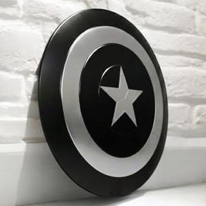 May include: A black and silver Captain America shield with a white star in the center. The shield is round with a black outer ring, a silver inner ring, and a white star in the center.