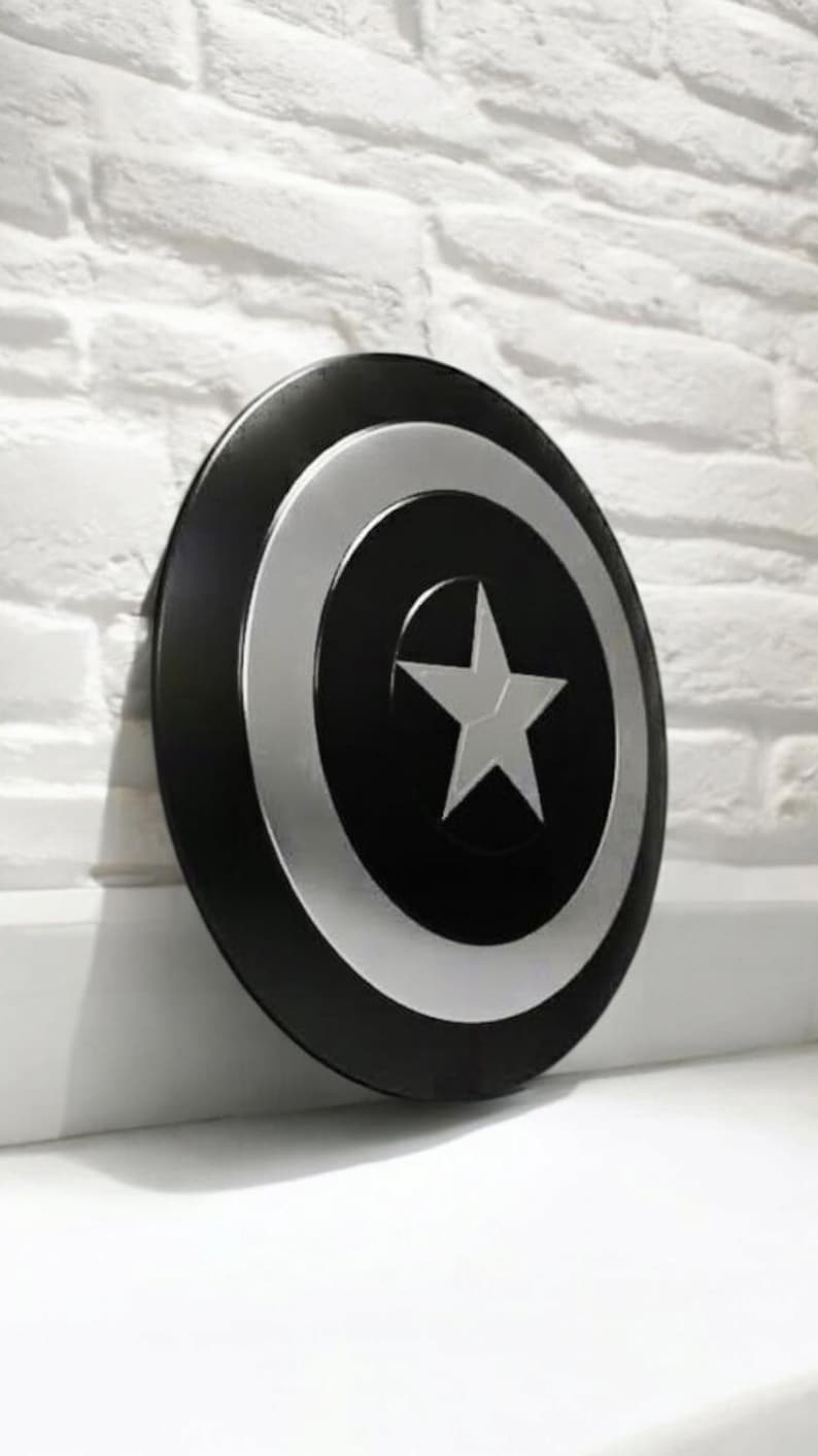 May include: A black and silver Captain America shield with a white star in the center. The shield is round with a black outer ring, a silver inner ring, and a white star in the center.