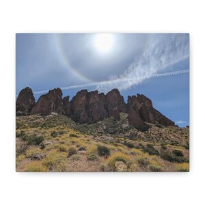 Könnte beinhalten: Leinwanddruck einer Wüstenlandschaft mit einer Bergkette unter einer hellen Sonne mit Halo-Effekt. Der Himmel ist blau mit ein paar Wolken und einer Kondensstrecke. Der Vordergrund zeigt trockene Vegetation.