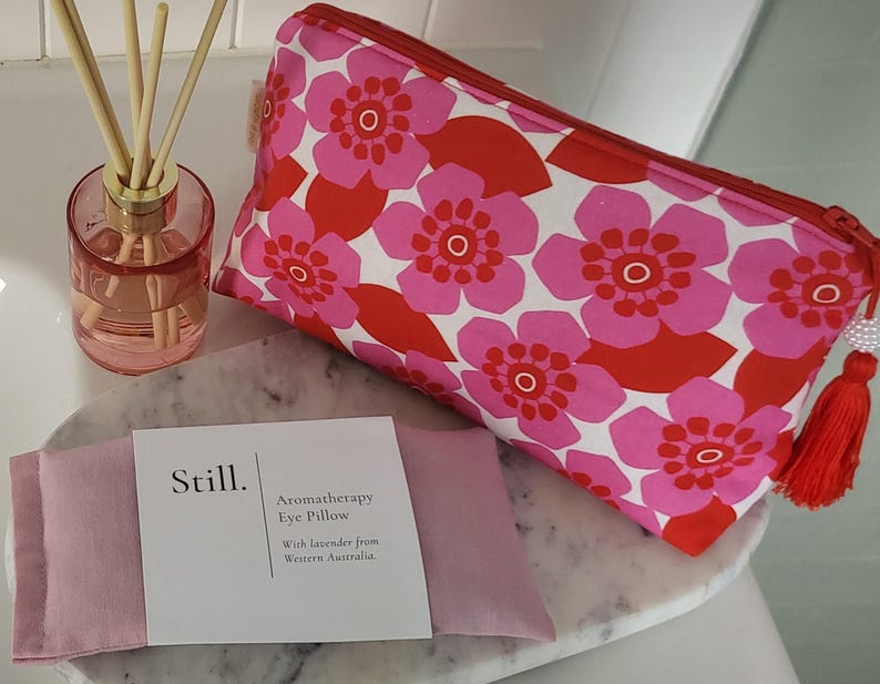 May include: A pink aromatherapy eye pillow with a white label that reads "Still. Aromatherapy Eye Pillow With lavender from Western Australia." A pink floral cosmetic bag and a pink glass diffuser with reeds are also in the image.