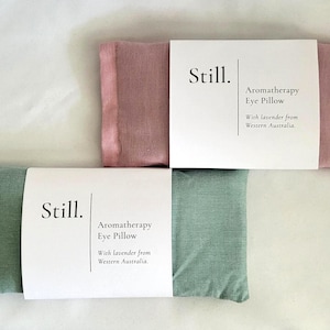 May include: Two aromatherapy eye pillows, one dusty rose and one sage green, are displayed. Each pillow is rectangular and has a white paper band with the word "Still" and the text "Aromatherapy Eye Pillow With lavender from Western Australia."