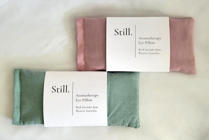 May include: Two aromatherapy eye pillows, one dusty rose and one sage green, are displayed. Each pillow is rectangular and has a white paper band with the word "Still" and the text "Aromatherapy Eye Pillow With lavender from Western Australia."